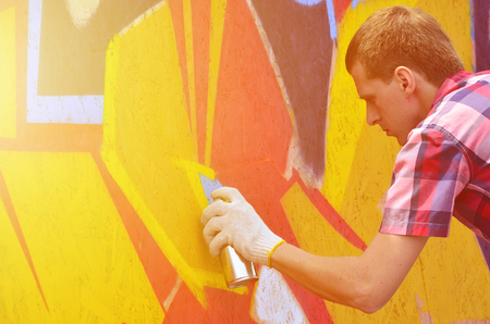 A young red-haired graffiti artist paints a new graffiti on the wall. Photo of the process of drawing a graffiti on a wall close-up. The concept of street art and illegal vandalismの写真素材