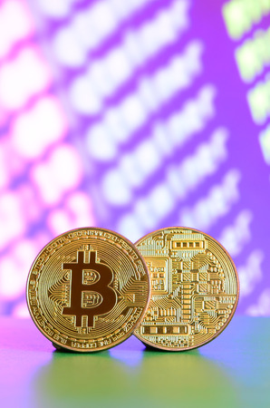 Two gold bitcoins lie on the green surface on the background of the display, which shows the process of mining the crypto currencyの写真素材