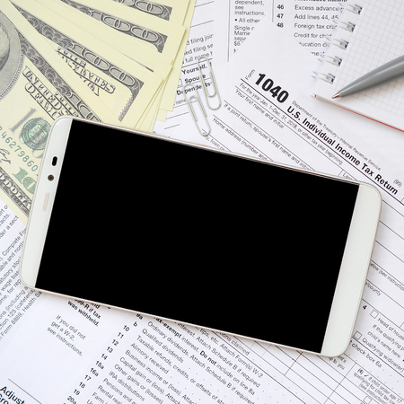 Composition of items lying on the 1040 tax form. Dollar bills, pen, smartphone, paper clip and notepad. Clean black screen smartphone for text placementの写真素材