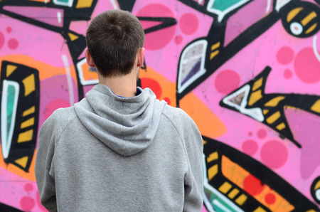 A young graffiti artist in a gray hoodie looks at the wall with his graffiti in pink and green colors on a wall in rainy weather. Street art conceptの写真素材