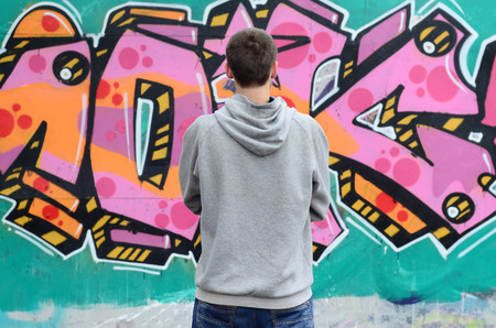 A young graffiti artist in a gray hoodie looks at the wall with his graffiti in pink and green colors on a wall in rainy weather. Street art conceptの写真素材