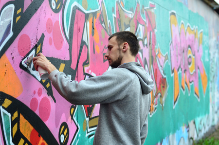 A young guy in a gray hoodie paints graffiti in pink and green colors on a wall in rainy weatherの写真素材