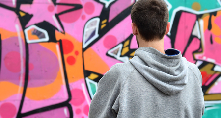A young graffiti artist in a gray hoodie looks at the wall with his graffiti in pink and green colors on a wall in rainy weather. Street art conceptの写真素材