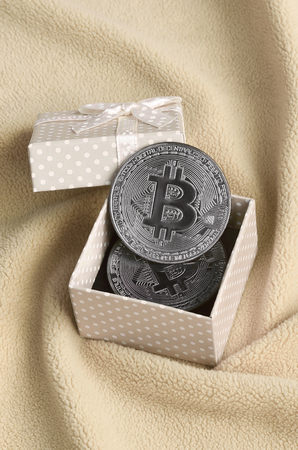 The silver bitcoin lies in a small orange gift box with a small bow on a blanket made of soft and fluffy light orange fleece fabric with a large number of relief foldsの写真素材