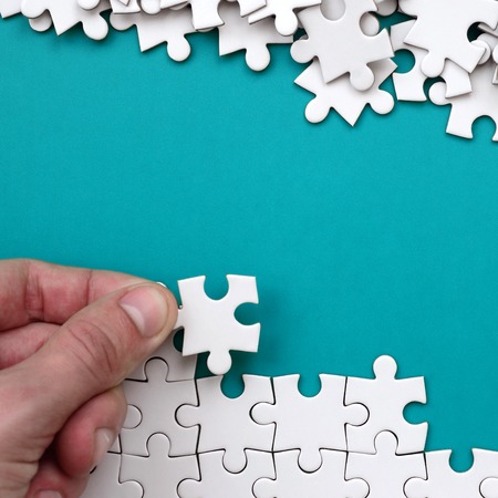 The hand folds a white jigsaw puzzle and a pile of uncombed puzzle pieces lies against the background of the blue surface. Texture photo with space for textの写真素材