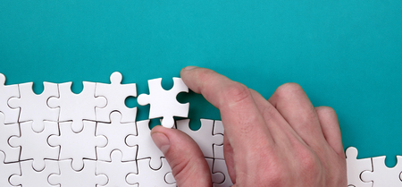 The hand folds a white jigsaw puzzle against the background of the blue surface. Texture photo with...