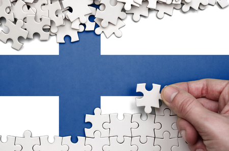 Finland flag  is depicted on a table on which the human hand folds a puzzle of white color.の写真素材