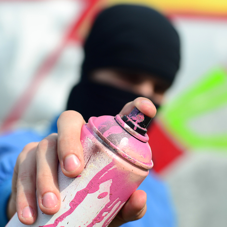 A young graffiti artist in a blue jacket and black mask is holding a can of paint in front of him against a background of colored graffiti drawing. Street art and vandalism concept.の写真素材