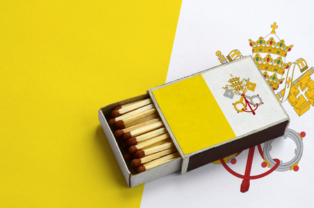 Vatican City State flag  is shown in an open matchbox, which is filled with matches and lies on a large flag.の写真素材