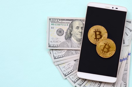 Bitcoins lies on a smartphone and hundred dollar bills on a light blue background.の写真素材
