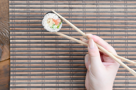 A hand with chopsticks holds a sushi roll on a bamboo straw serwing mat background. Traditional Asian food.の写真素材