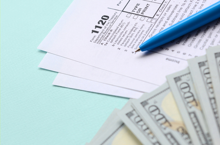 1120 tax form lies near hundred dollar bills and blue pen on a light blue background. US Corporation income tax return.の写真素材