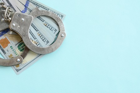 Silver police handcuffs and hundred dollar bills lies on light blue background.