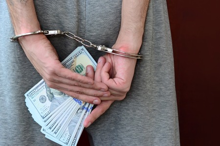An arrested guy in gray pants with handcuffed hands holds a huge amount of dollar bills. Back view.