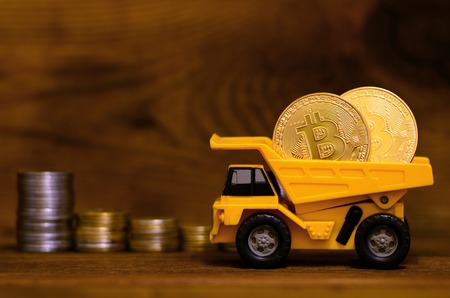 Yellow toy dumper loaded with shiny golden bitcoins is standing next to stacks of coins of various currencies on a wooden background. The advantage of digital currency versus physical moneyの写真素材