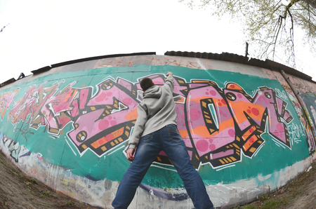 A young guy in a gray hoodie paints graffiti in pink and green colors on a wall in rainy weather. Fisheye shotの写真素材