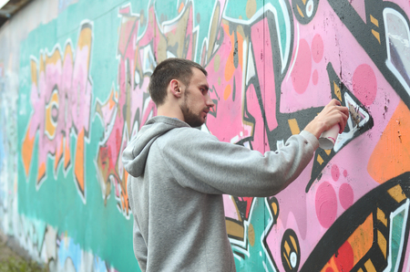 A young guy in a gray hoodie paints graffiti in pink and green colors on a wall in rainy weatherの写真素材