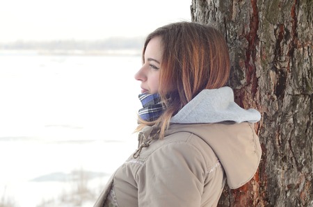 A young Caucasian girl in a brown coat staring into the distance on the horizon line between the sky and the frozen lake in winterの写真素材