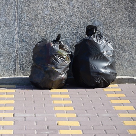 Two black garbage bags on tiled street floor at concrete fence in city. Pollution trash