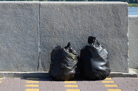 Two black garbage bags on tiled street floor at concrete fence in city. Pollution trashの写真素材