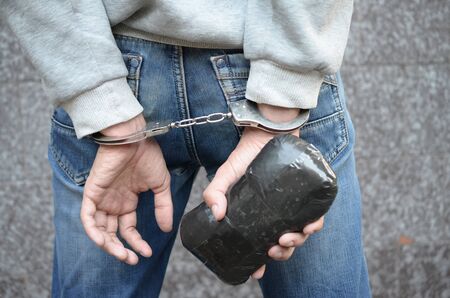 Arrested drug dealer in handcuffs with big heroin drug package on dark wall background. Illegal drug...