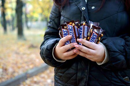 KHARKOV, UKRAINE - OCTOBER 8, 2019: A young caucasian woman shows many Snickers chocolate bars in autumn park. Snickers chocolate manufactured by Marsのeditorial素材