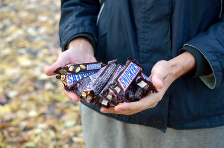KHARKOV, UKRAINE - OCTOBER 8, 2019: A young caucasian man shows many Snickers chocolate bars in autumn park. Snickers chocolate manufactured by Marsのeditorial素材