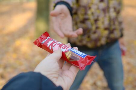 KHARKOV, UKRAINE - OCTOBER 21, 2019: A male hand passes the girl a Kit kat chocolate bar in an autumn park. The manifestation of kindness, treating with sweetsのeditorial素材
