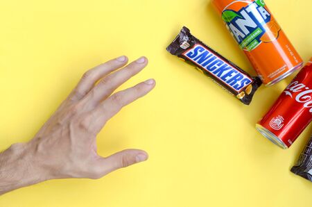 KHARKOV, UKRAINE - OCTOBER 2, 2019: Male hand reaches for Snickers chocolate bar in brown wrapping, Coca Cola and Fanta tin can on yellow pastel background. Famous drink and chocolate productのeditorial素材