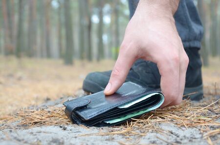 Male hand picking up a lost wallet from a ground in autumn fir wood path. The concept of finding a...