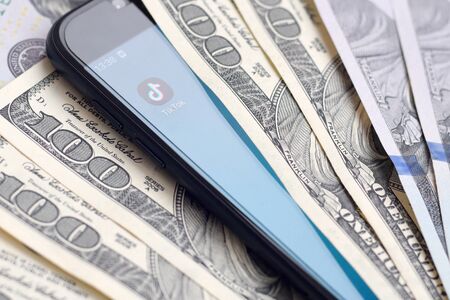 KHARKOV, UKRAINE - FEBRUARY 6, 2020: Smartphone screen with Tiktok app and lot of hundred dollar bills. Business and social networking conceptのeditorial素材