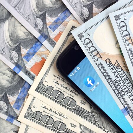 KHARKOV, UKRAINE - FEBRUARY 6, 2020: Smartphone screen with Facebook app and lot of hundred dollar bills. Business and social networking conceptのeditorial素材