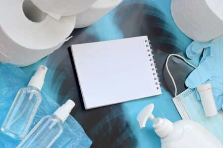 Set of items for Covid-19 quarantine. Empty notepad page and toilet paper, rubber disposable gloves with surgical face mask and hand sanitizer with liquid soap bottle on human chest x-ray filmの写真素材