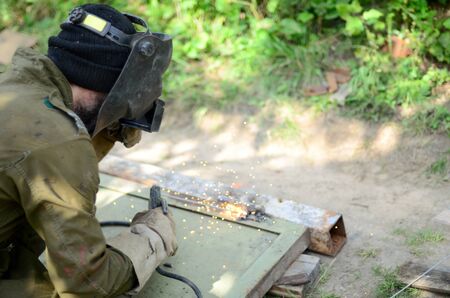 Old man welder in brown uniform, welding mask and welders leathers, weld metal door with arc welding machine outdoors, blue sparks fly to the sidesの写真素材