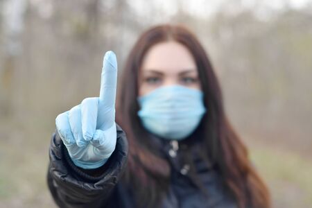 Portrait of young brunette woman in blue protective mask and rubber gloves shows attention gesture outdoors in spring wood. Concept of protective goods usage during quarantineの写真素材