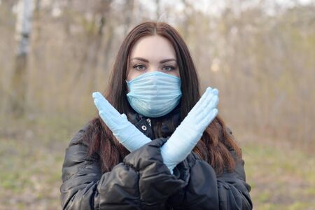 Portrait of young brunette woman in blue protective mask and rubber gloves shows stop gesture outdoors in spring wood. Concept of protective goods usage during quarantineの写真素材