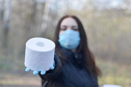 Covidiot concept. Young woman in protective mask gives roll of toilet paper outdoors in spring wood....