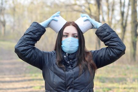 Covidiot concept. Young woman in protective mask holds many rolls of toilet paper outdoors in spring wood. Panic buying during quarantineの写真素材