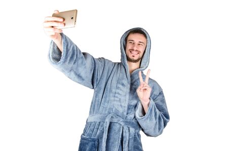 Portrait of young caucasian bearded man in blue bathrobe shows victory gesture and makes selfie isolated on white background. Happy emotion conceptの写真素材