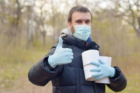 Covidiot concept. Young man in protective mask holds many rolls of toilet paper and shows thumb up outdoors in spring wood. Panic buying during quarantineの写真素材