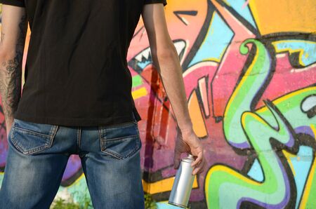 Young graffiti artist with backpack and gas mask on his neck paints colorful graffiti in pink tones on brick wall. Street art and contemporary painting process. Entertainment in youth subcultureの写真素材