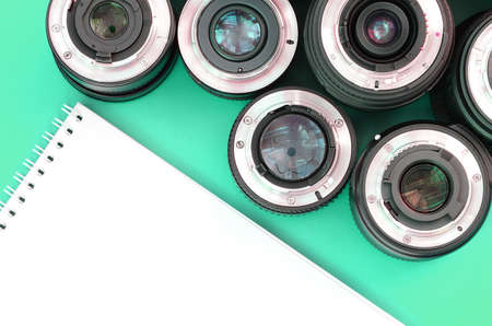Several photographic lenses and white notebook lie on a bright turquoise background. Space for text