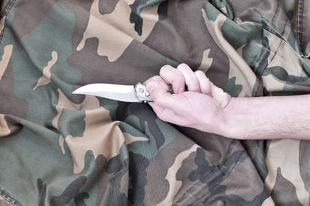 Hand with small knife on camouflage fabric background. The concept of using a knife in melee tactics...
