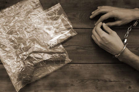 Handcuffed hands of criminal suspect on wooden table and crime scene evidence in transparent plastic packs for investigation. Handgun and jackknifeの写真素材