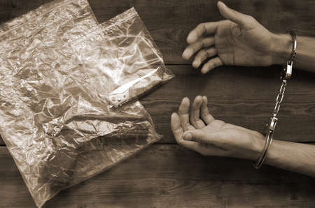 Handcuffed hands of criminal suspect on wooden table and crime scene evidence in transparent plastic packs for investigation. Handgun and jackknifeの写真素材