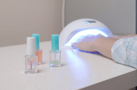 Three colorless transparent nail polish bottles in background of female hands in gel uv led nail white lamp for drying manicure. Beauty care women procedures in salon or home on gray backgroundの写真素材