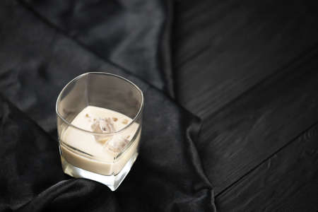 Glass of Irish cream liqueur with ice cubes on dark fabric and wooden bar table background. Homemade luxury elite alcohol cocktail. Place for your textの写真素材