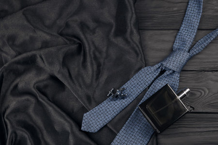 A bottle of mens cologne and cufflinks with blue tie lie on a black luxury fabric background on a wooden table. Mens classic accessories. Shallow DOFの写真素材