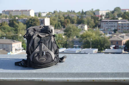 Black backpack lies on metal border of residental multistorey building rooftop in sunny weather outdoorsの写真素材
