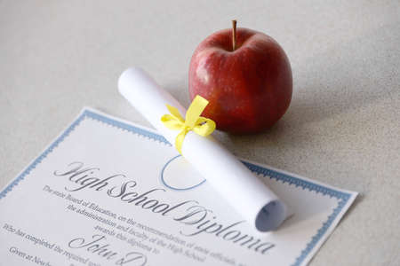 A high school diploma lies on table with small scroll and red apple. Education documents close up